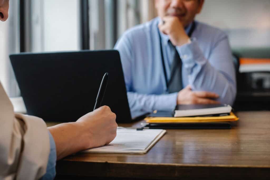 An image of a person writing notes on a clipboard and another man behind a computer. One is drafting his resume manually and the other has already completed his resume with an AI Resume Builder. He is waiting and pondering why the other person didn't use an AI Resume Builder.